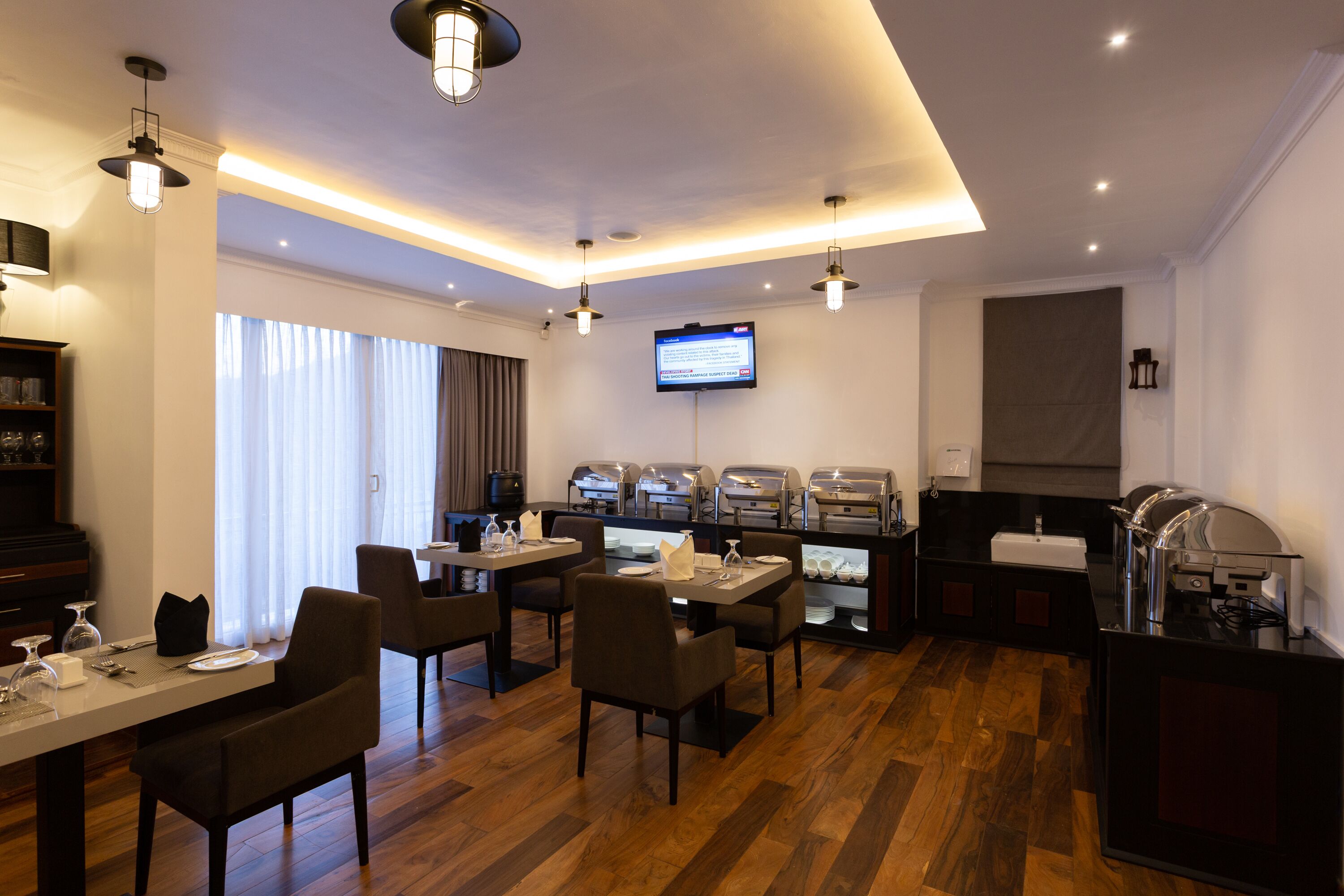 A modern hotel dining area with wooden flooring, contemporary furniture, a buffet setup, and a flat-screen TV mounted on the wall.