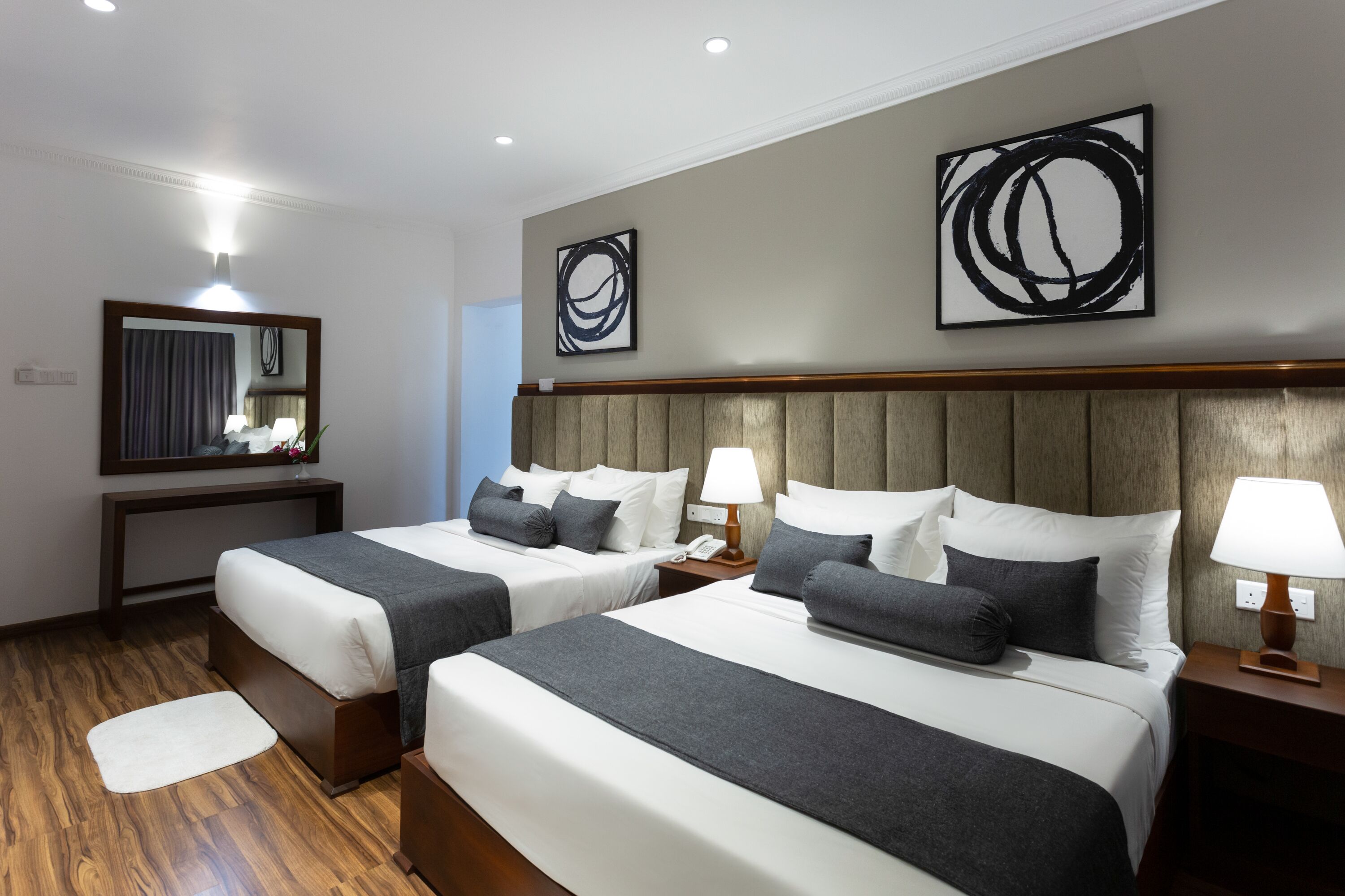 A hotel room with two beds, a wooden headboard, bedside lamps, a mirror, and abstract wall art.