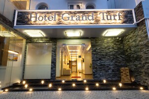 La entrada del Hotel Grand Turf, con fachada de piedra, puerta de cristal y alfombra roja que conduce a la entrada.