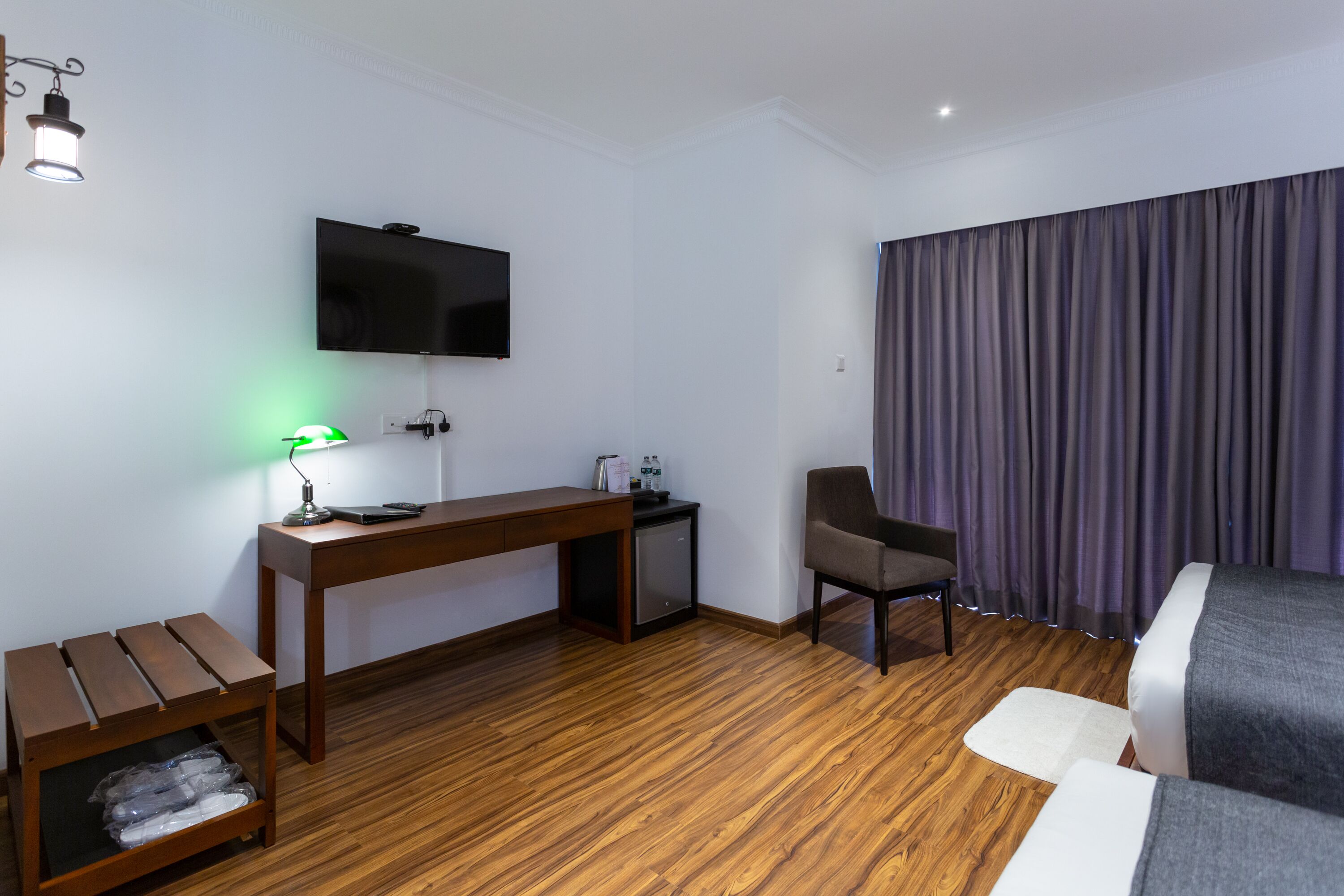 A hotel room with a wooden floor, a desk with a lamp, a chair, a bed, a TV mounted on the wall, and a window with curtains.
