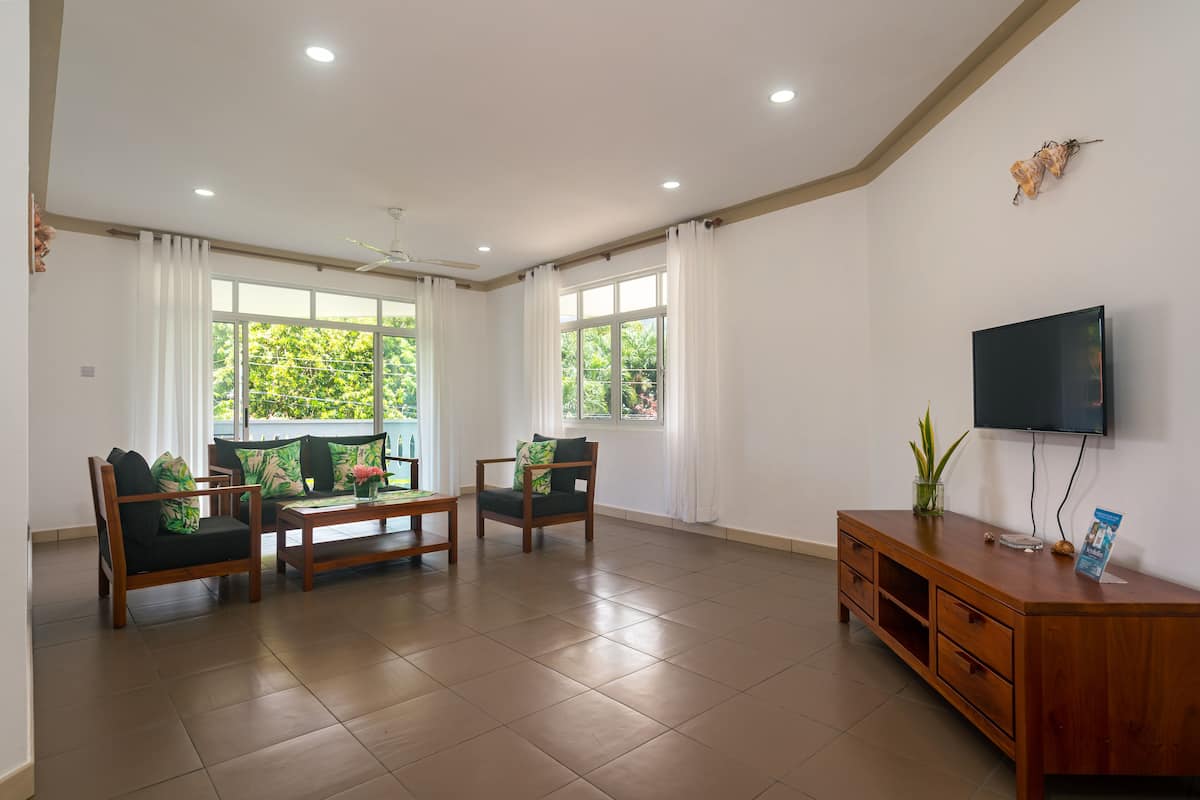 A spacious living room with a flat-screen TV, wooden furniture, and large windows with curtains.