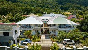 A large, two-story building with a central entrance, surrounded by lush greenery and multiple vehicles parked in front. - 340 Mountain View Apartments (Mahé Island)