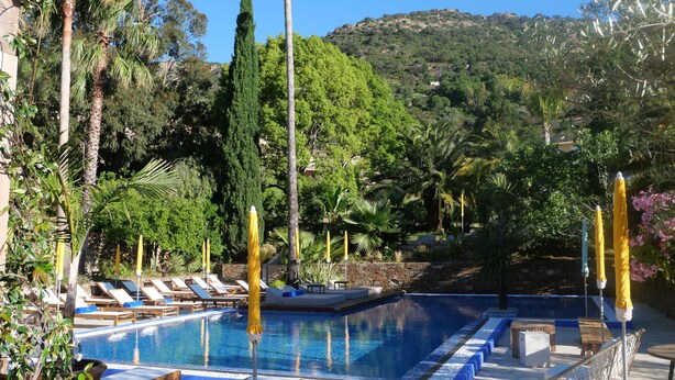 Un espace piscine doté de chaises longues et de parasols, entouré d’arbres et avec une chaîne de montagnes en toile de fond.