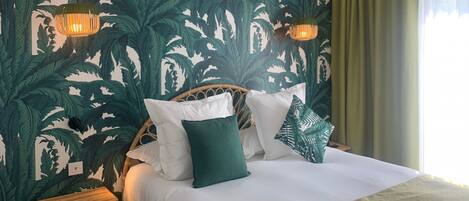 Una cama con ropa blanca, almohadas verdes y blancas, y un papel tapiz con motivos de hojas tropicales.