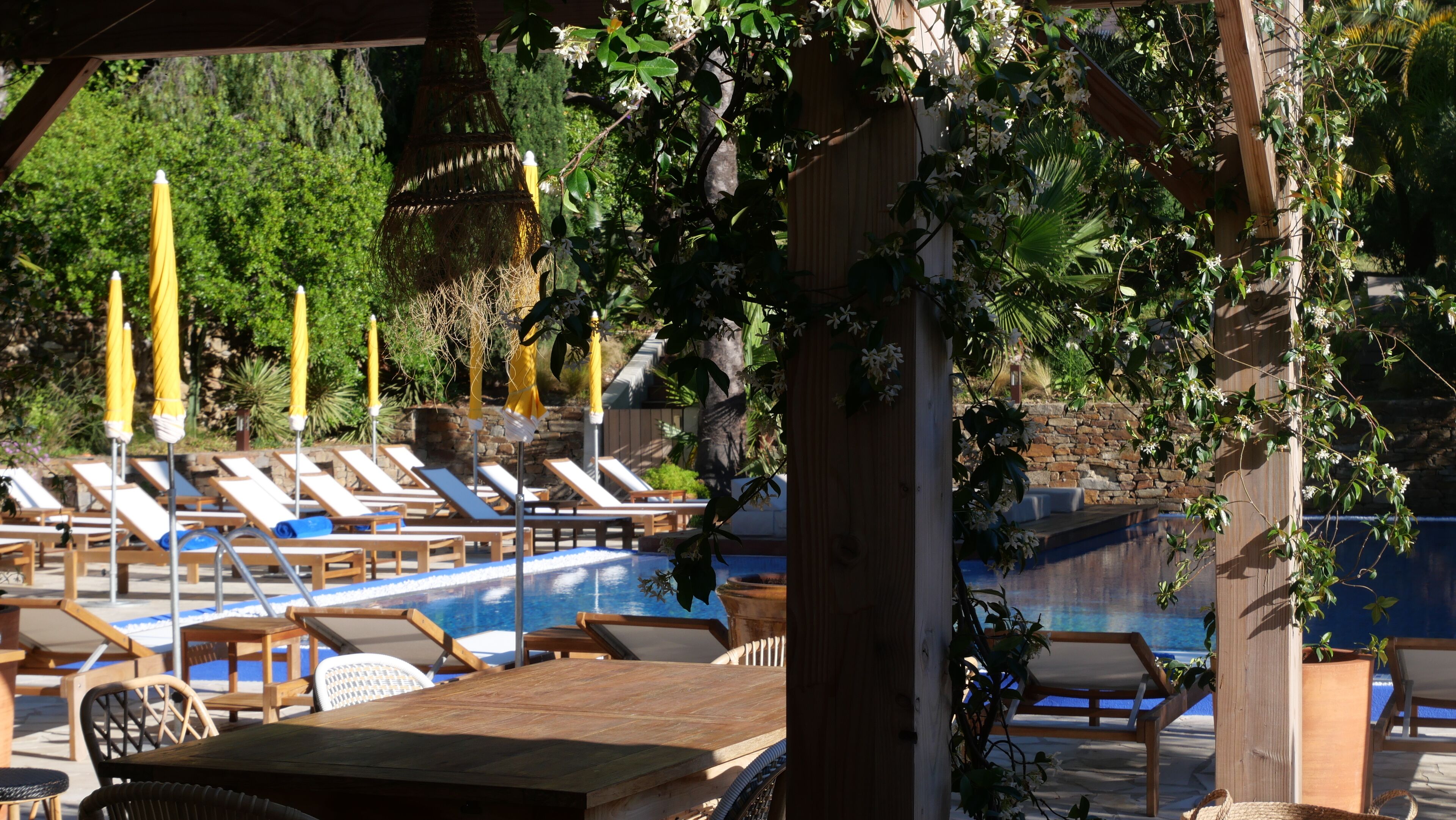 Een zwembadterras met ligbedden, houten tafels en gele parasols.
