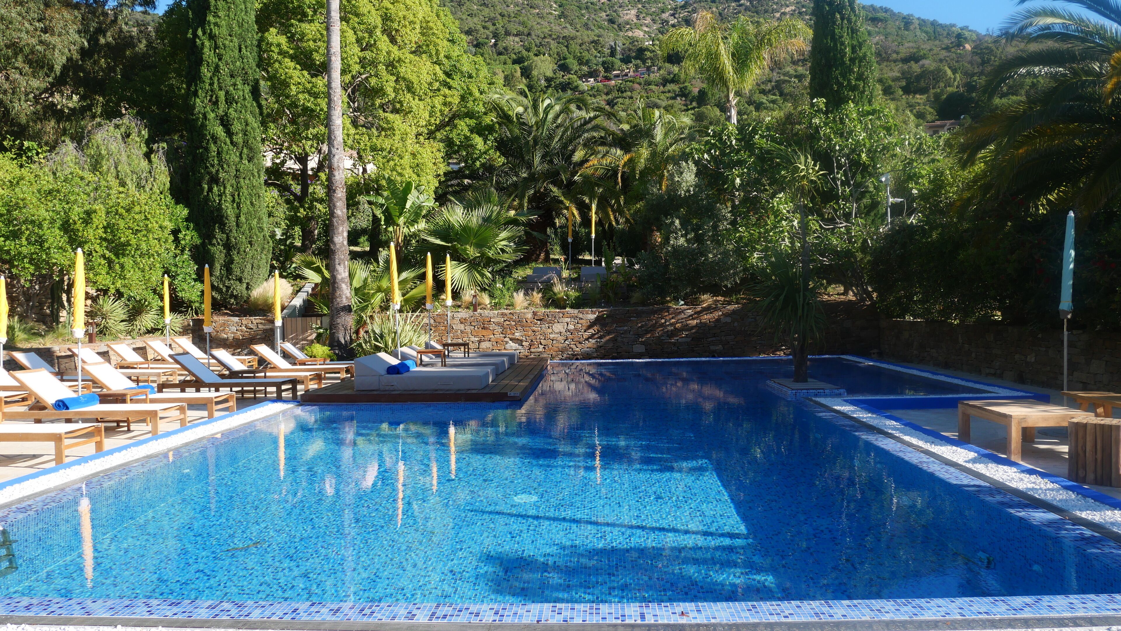 Una piscina azul cristalina con sillas de descanso y sombrillas, rodeada de exuberante vegetación y muros de piedra.