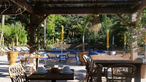 Un espace piscine extérieur avec des chaises longues, une table à manger pour deux personnes et une pergola ornée de plantes suspendues.