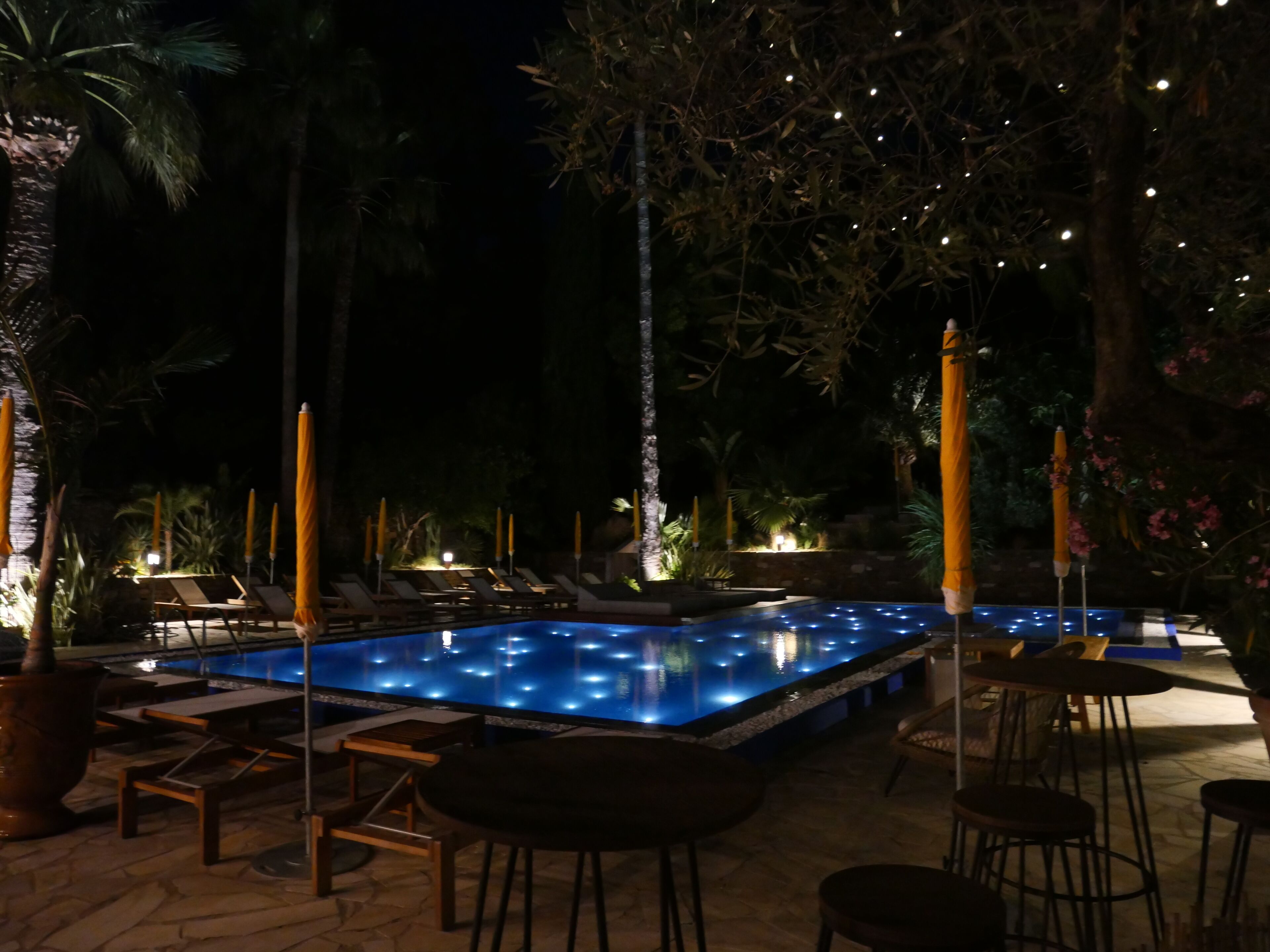 Un área de piscina nocturna con sillones de descanso y mesas, rodeada de palmeras e iluminada con luces colgantes.