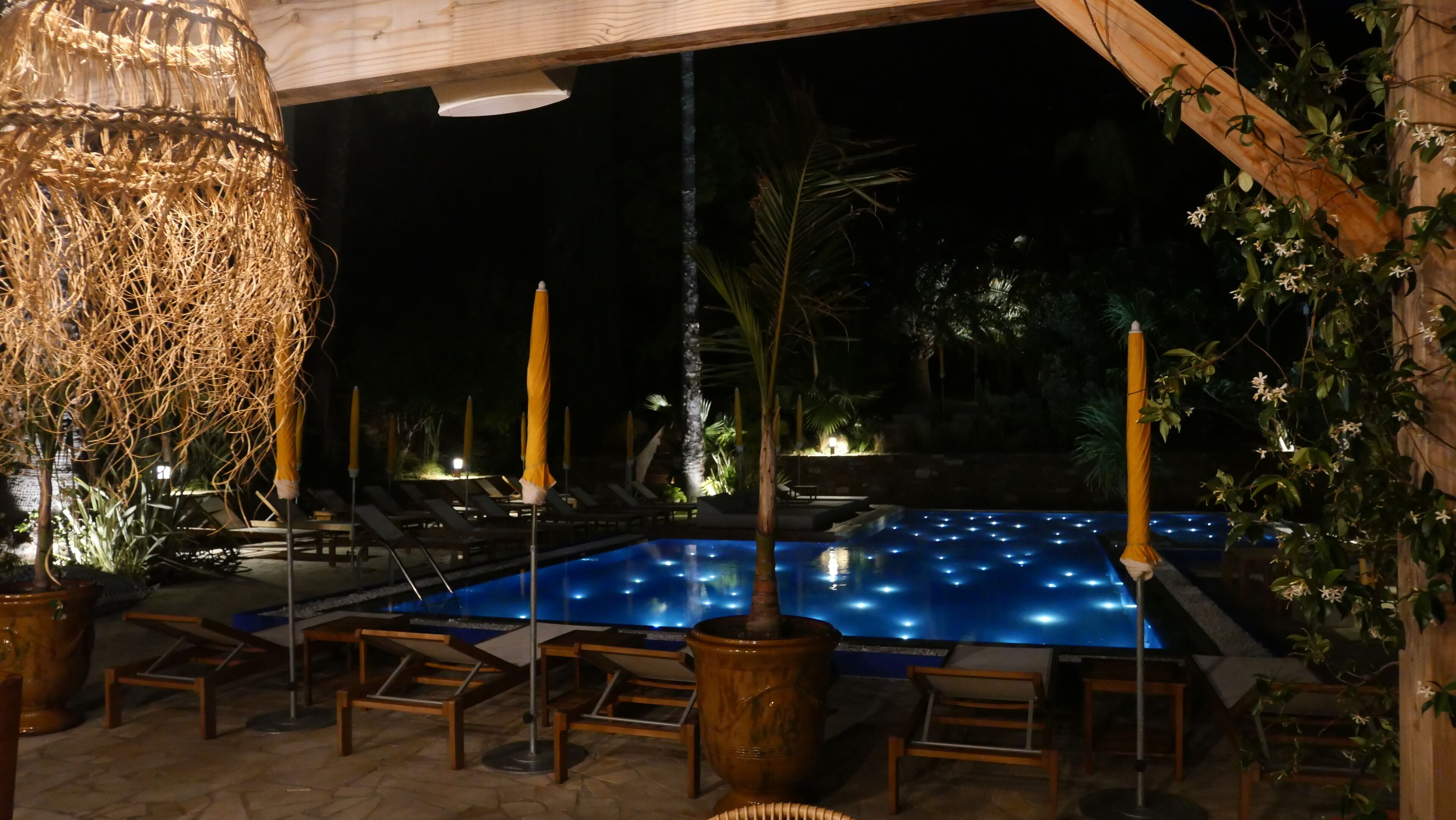 Un área de piscina nocturna con sillones reclinables y una lámpara colgante decorativa.