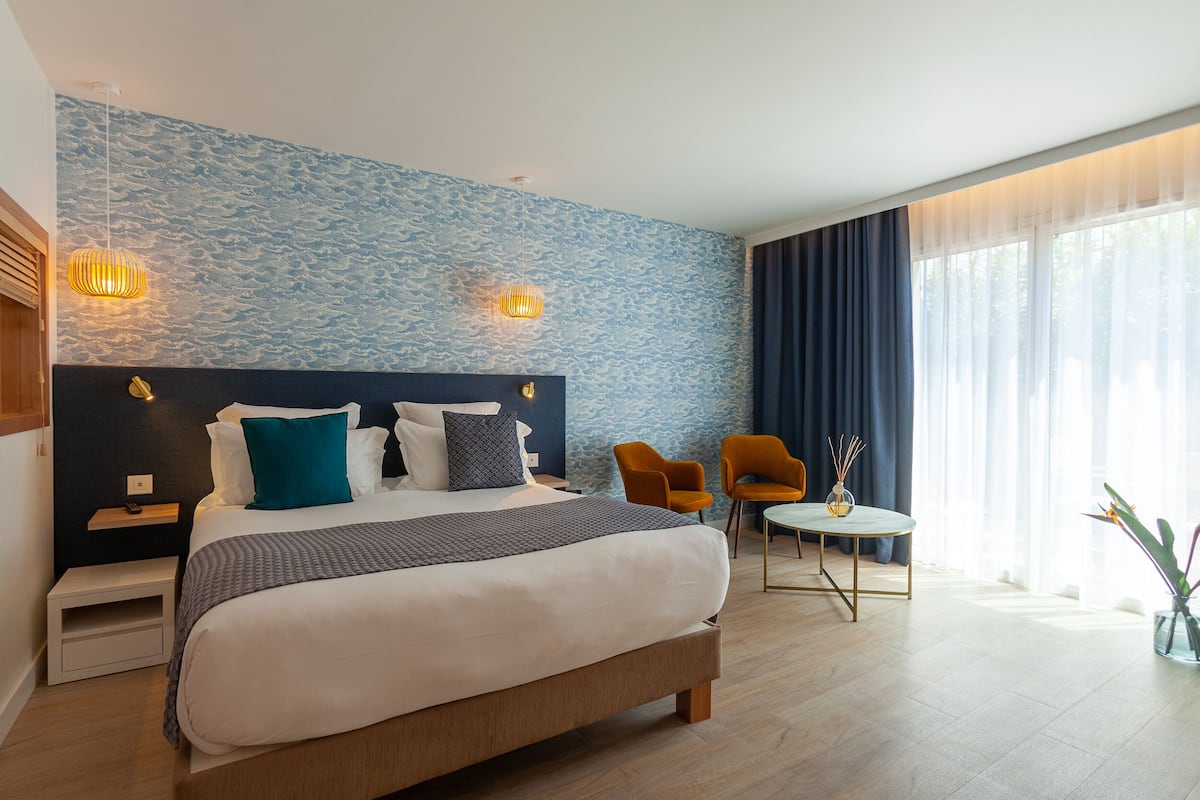 Een moderne hotelkamer met een groot bed, twee nachtkastjes, een kleine tafel met stoelen en een groot raam met gordijnen.