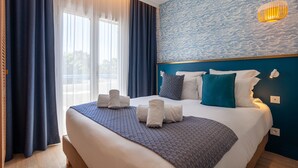 Un lit bien fait, agrémenté de serviettes, d’une couverture à motifs et d’une tête de lit bleue, dans une chambre d’hôtel dotée d’une grande fenêtre et de rideaux.