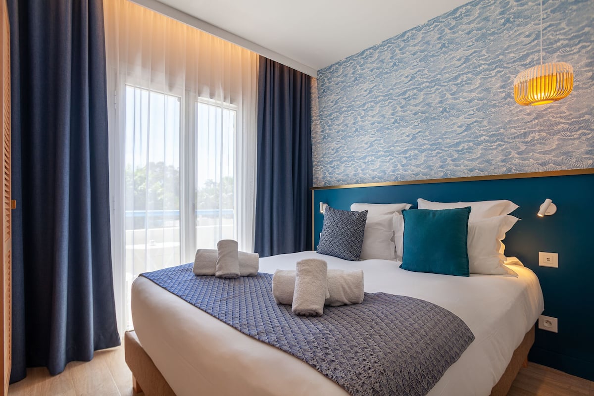 Een net opgemaakt bed met handdoeken, een geborduurde deken en een blauw hoofdeinde in een hotelkamer met een groot raam en gordijnen.
