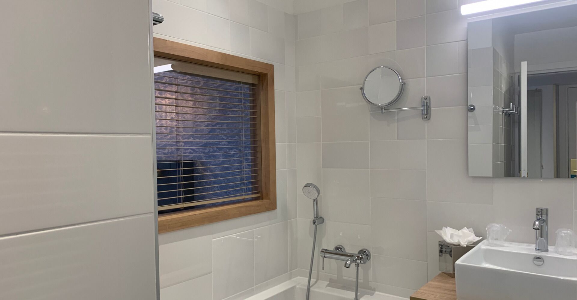 A modern bathroom with white tiles, a wooden-framed window, a shower, a sink, and a mirror.