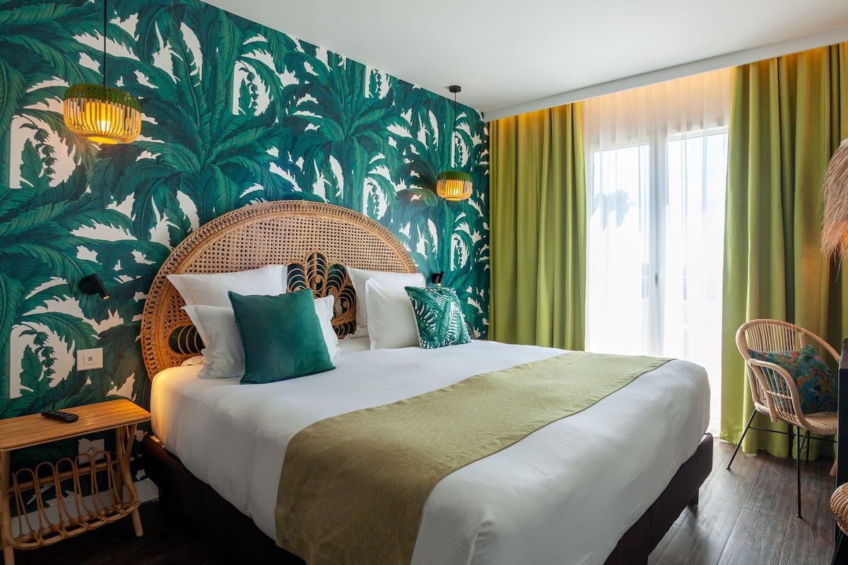 Een hotelkamer met een bed, een rieten stoel, een klein tafeltje en een raam met gordijnen.