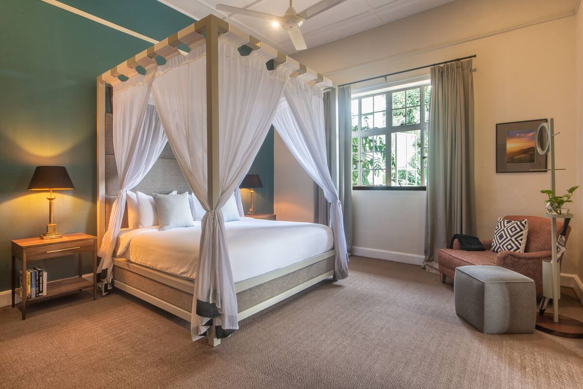 A four-poster bed with a canopy, a bedside table with a lamp, a reading chair, and a small ottoman.