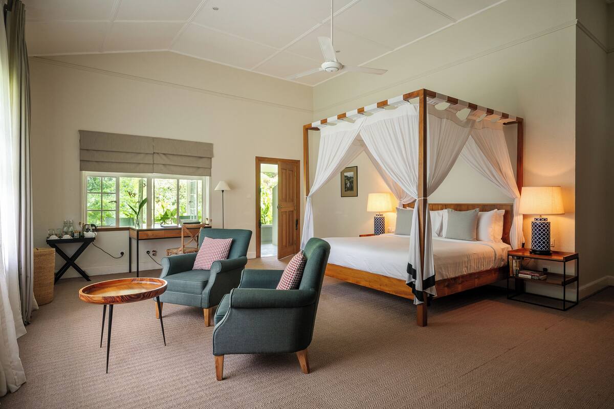 A four-poster bed, two armchairs, a small table, and a bedside table with lamps in a well-lit room with large windows.