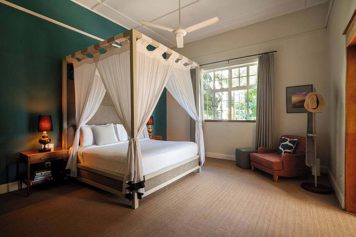 A four-poster bed with a canopy, a nightstand with a lamp, a chair, and a large window with curtains.