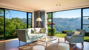 Uma sala de estar moderna com sofá, poltrona e mesa de centro, grandes janelas com vista para vegetação e colinas.