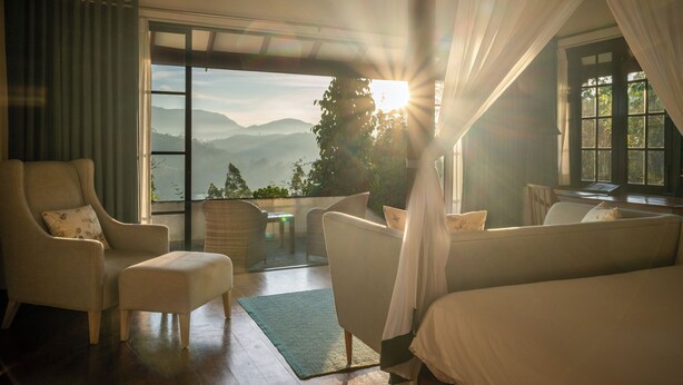 Quarto com uma cama grande, uma cadeira, uma mesinha e vista para as montanhas pela janela.