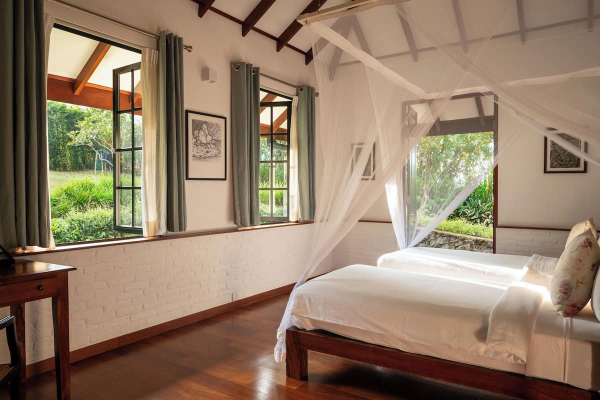 Ein Schlafzimmer mit einem hölzernen Bett, weißer Bettwäsche und Blick auf Grün hinter großen Fenstern.