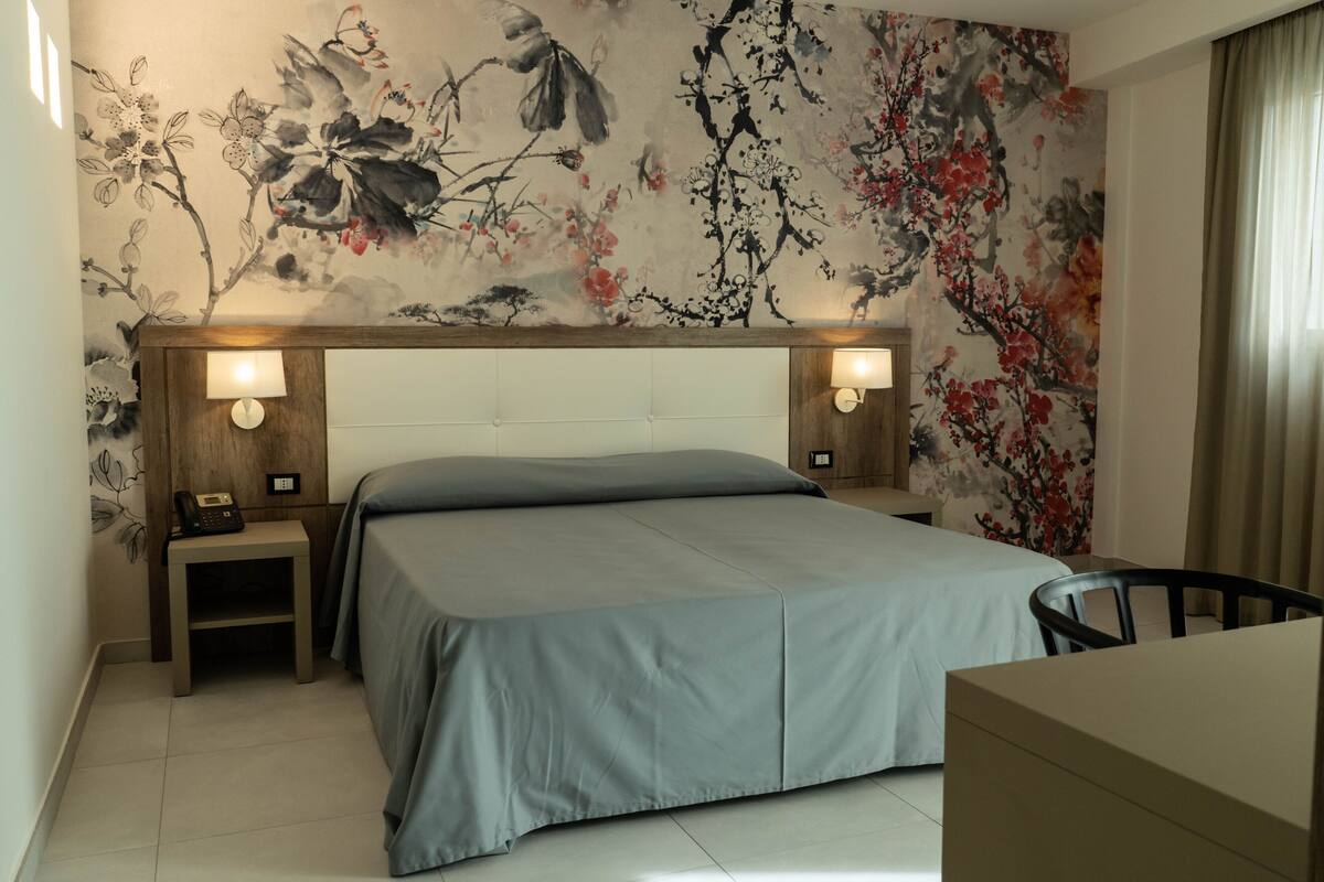 Hotelkamer met een groot bed, twee nachtkastjes met lampen, een bureau met stoel en een bloemen behang.