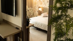 A hotel room with a bed, a bedside lamp, a TV mounted on the wall, a potted plant, and a wooden door frame. - Hotel Pontemare (Palermo)