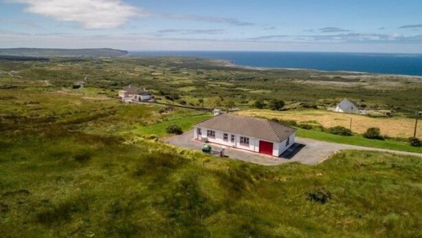 A white building with a red door, surrounded by green fields and a view of the sea. - Atlantic Rose Cottage (Lisdoonvarna)