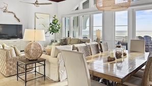 A spacious living room with a large wooden dining table, comfortable seating, a television, and a view of the ocean through large windows. - "Views from Above" | Beachfront | Community Heated Pool (Panama City Beach)