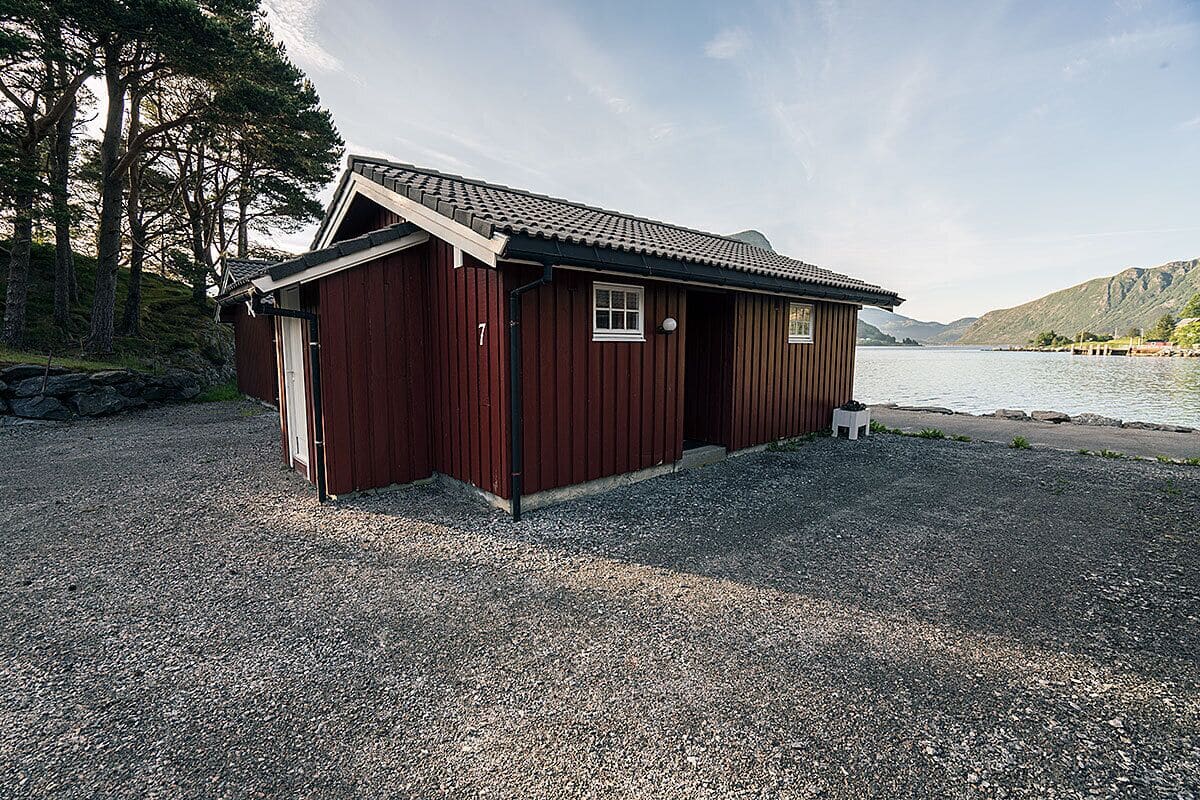 A small wooden cabin with a sloped roof, a door, and a window, situated on a gravel path with a lake and mountains in the background.