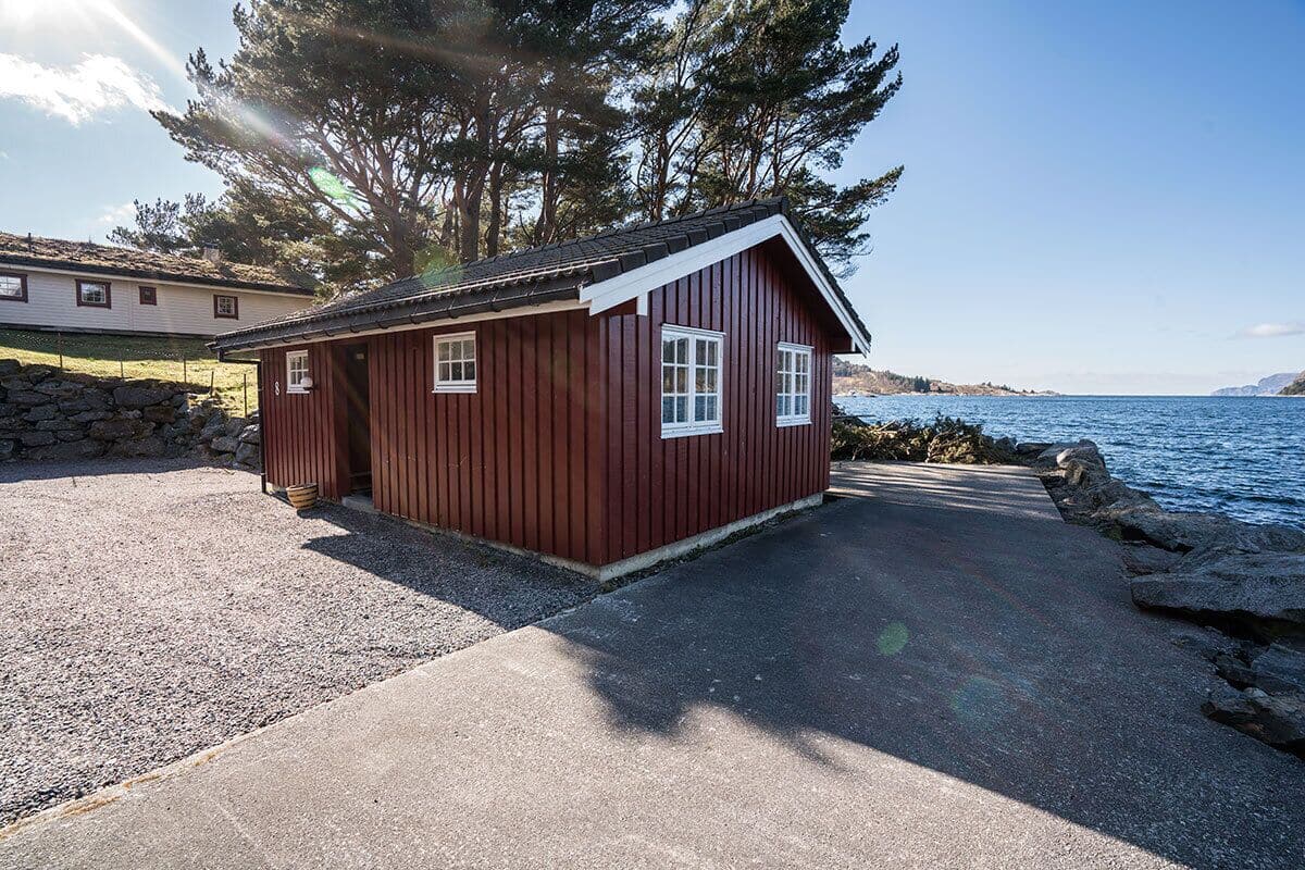 A small red wooden cabin with a gravel driveway and a stone wall, located near a body of water.