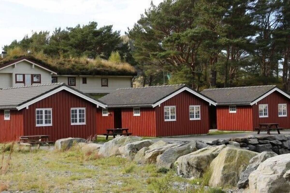 A row of red wooden cabins with white windows, a stone wall in the foreground, and a building with a green roof in the background.
