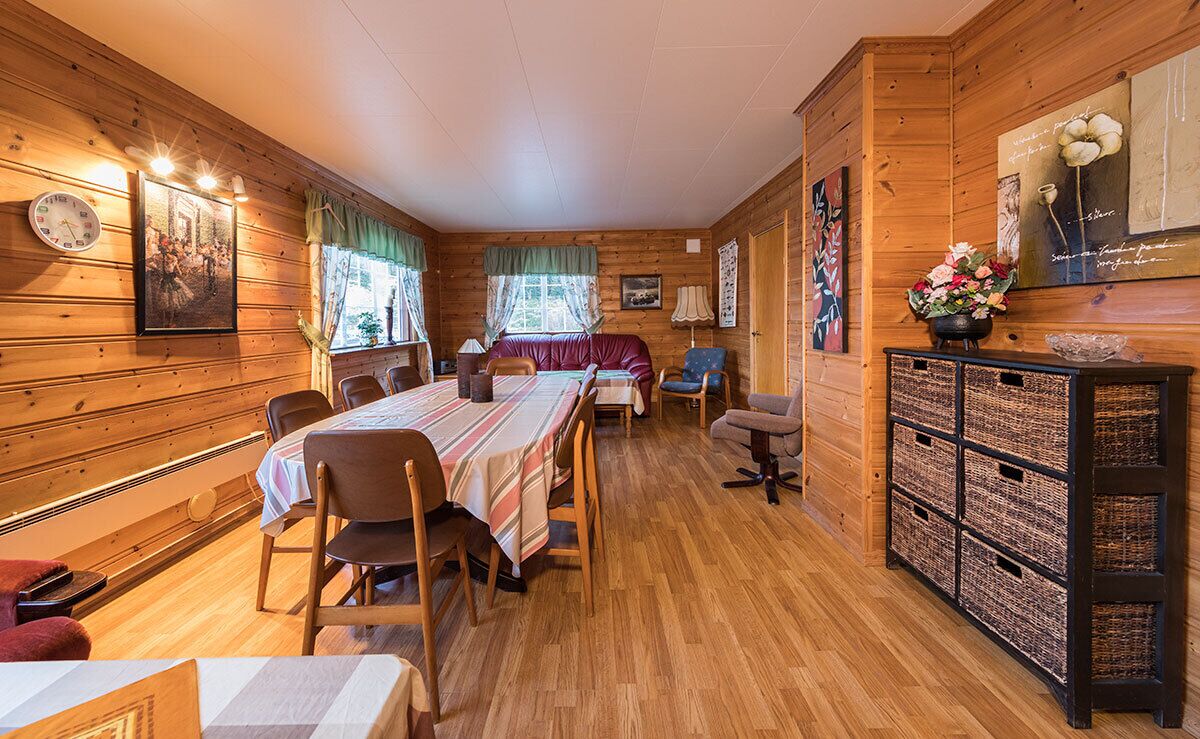 Ein Interieur einer Holzhütte mit Esstisch, Stühlen, einer Couch, einem kleinen Tisch mit Lampe und einem Schrank mit Körben.