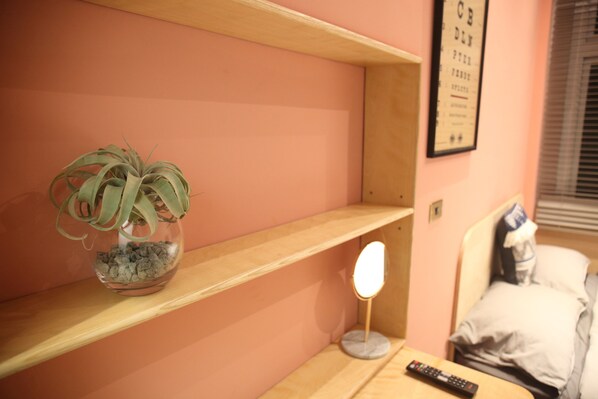 A room with a pink wall, a wooden shelf, a potted plant, a bedside table with a lamp, a remote control, and a bed with pillows.