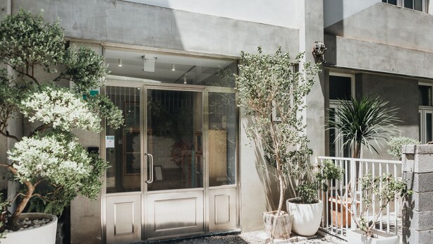 Entrada de un edificio con puerta de vidrio, plantas en macetas y una valla blanca.