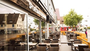 An outdoor cafe with wicker tables and chairs, a wooden facade, and a view of the street. - LOOK Hotel & Rooms (Crailsheim)