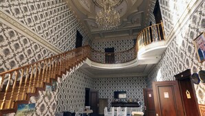 Una estancia imponente con papel tapiz ornamentado, un candelabro, una escalera de madera y un comedor con sillas blancas y una mesa.