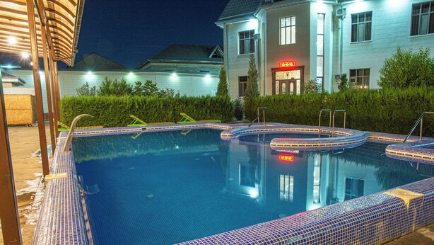 Una piscina con borde de azulejos y un jacuzzi, rodeados por una terraza de madera y un edificio con un cartel iluminado al fondo.