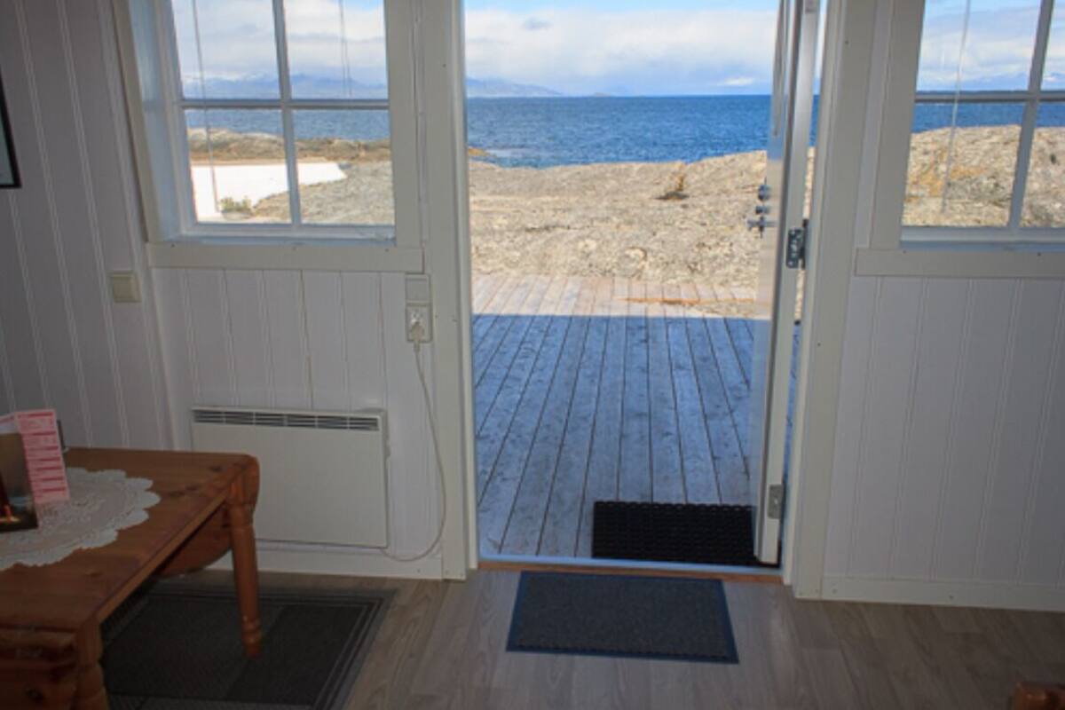 A room with a wooden floor, a table with a lace tablecloth, and a view of a beach through a door.