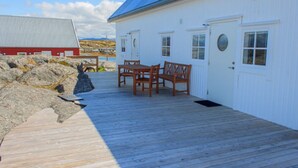 Une terrasse en bois mène à un bâtiment blanc surmonté d’un toit bleu, un bâtiment rouge se dressant à l’arrière-plan, et une côte rocheuse s’étend le long de la plage.