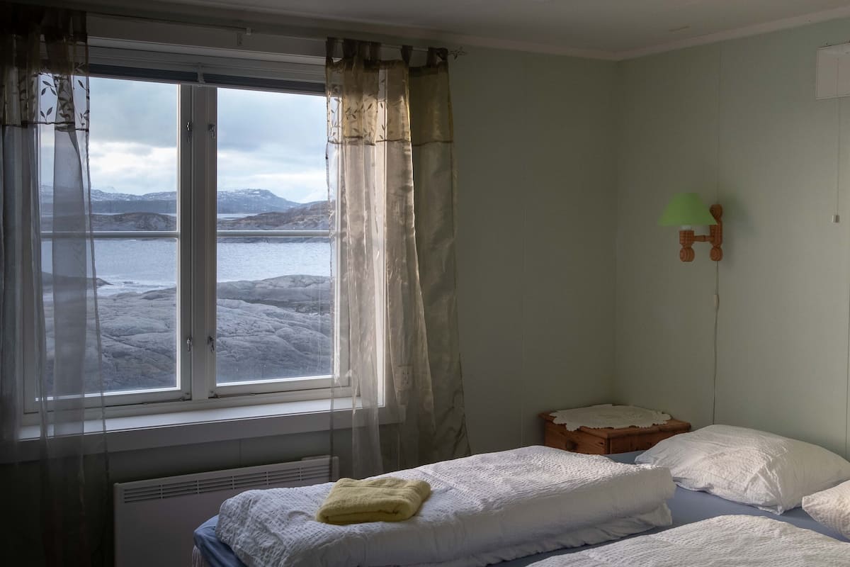 A room with two beds, a window with curtains, a radiator, and a wooden nightstand with a lamp.