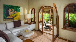 A room with a white sofa, a small round table, a painting, and a view of a garden through a wooden archway. - Villa Las Rocas (Cabo San Lucas)