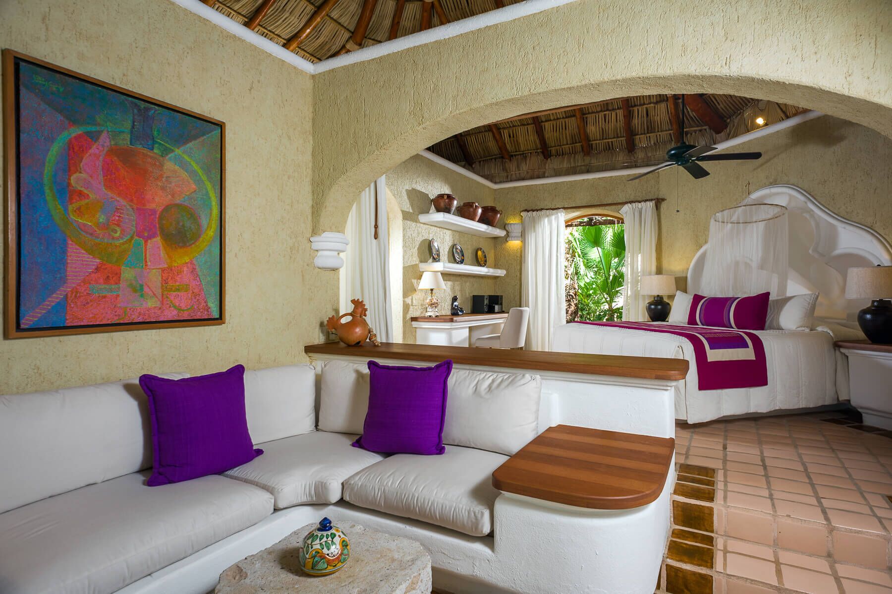 A cozy living area with a white sofa, purple cushions, a small wooden table, and a colorful abstract painting.