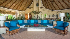 A spacious lounge area with a thatched roof, a large blue sofa, two wooden armchairs, a central coffee table, and a dining area in the background. - Villa Las Rocas (Cabo San Lucas)