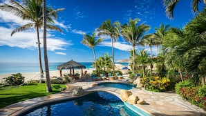 A pool area with a curved design, surrounded by palm trees and a beach view. - Villa Las Rocas (Cabo San Lucas)