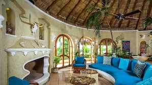 A tropical-style living room with a curved ceiling, a fireplace, a large sofa, and a ceiling fan. - Villa Las Rocas (Cabo San Lucas)