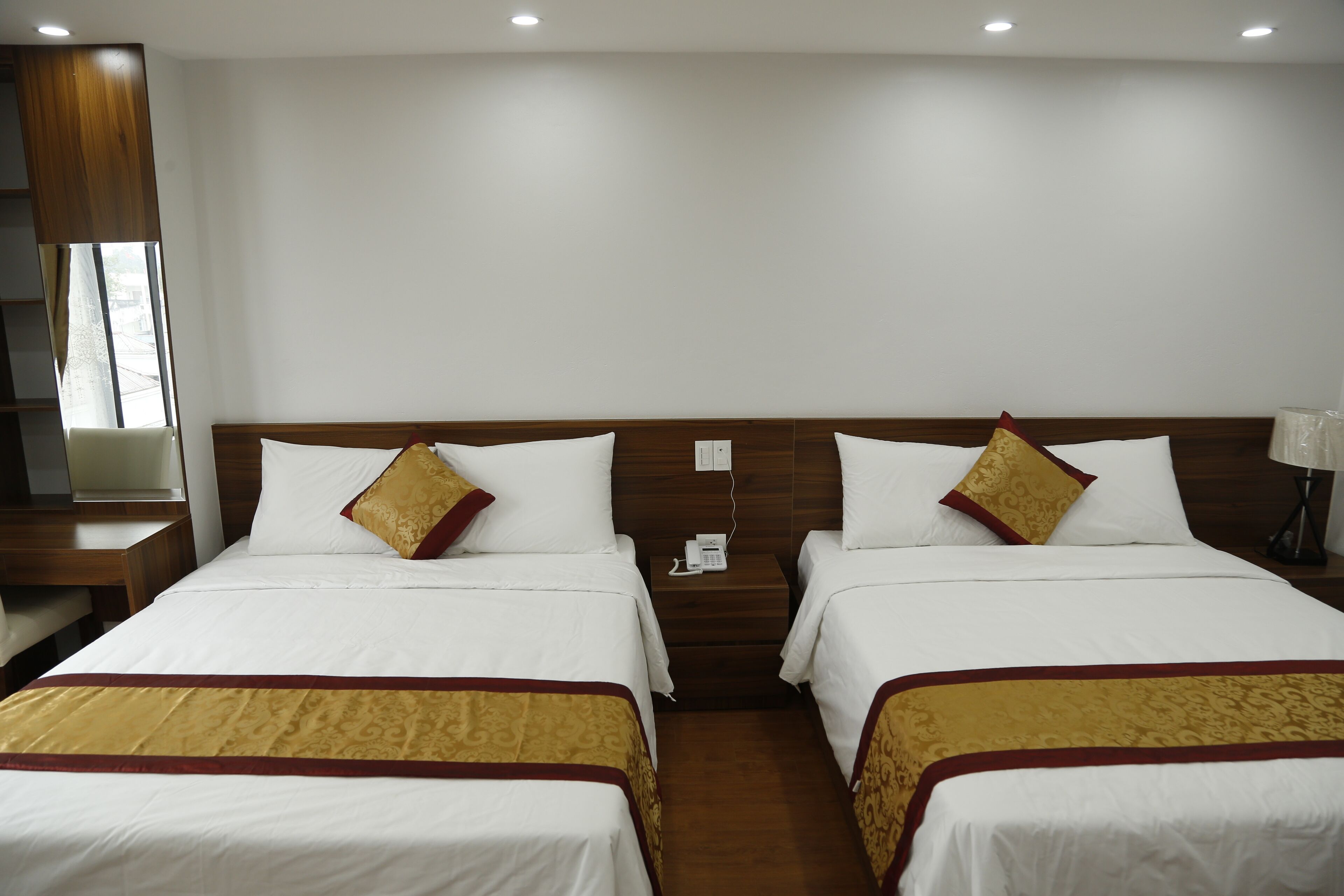 Habitación de hotel con dos camas, cada una con cabecero de madera, ropa de cama blanca y una manta con estampado dorado. Hay una mesita de noche con un teléfono y una lámpara entre las camas.