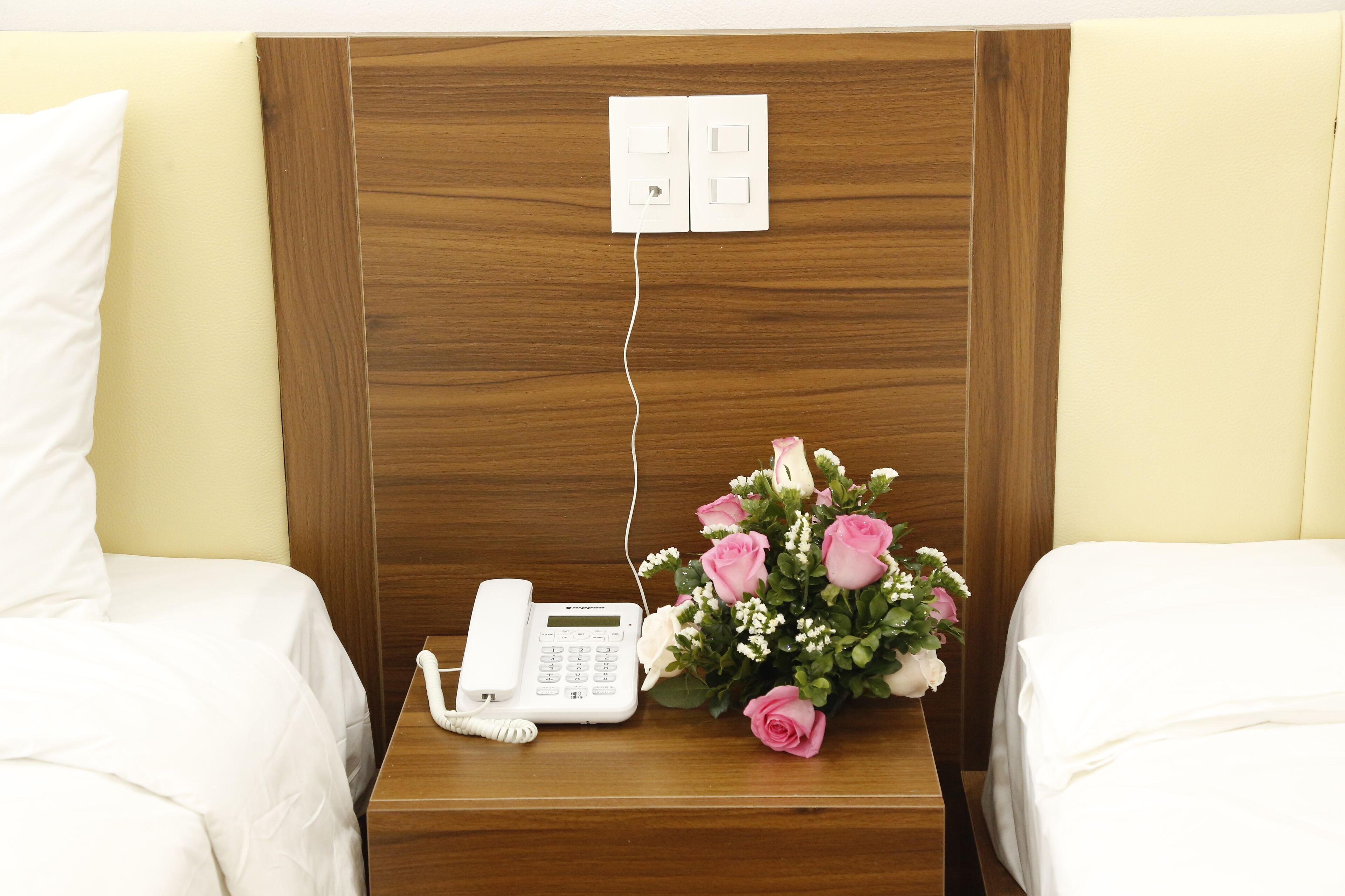 A hotel room with a wooden headboard, a bedside phone, and a bouquet of pink roses.