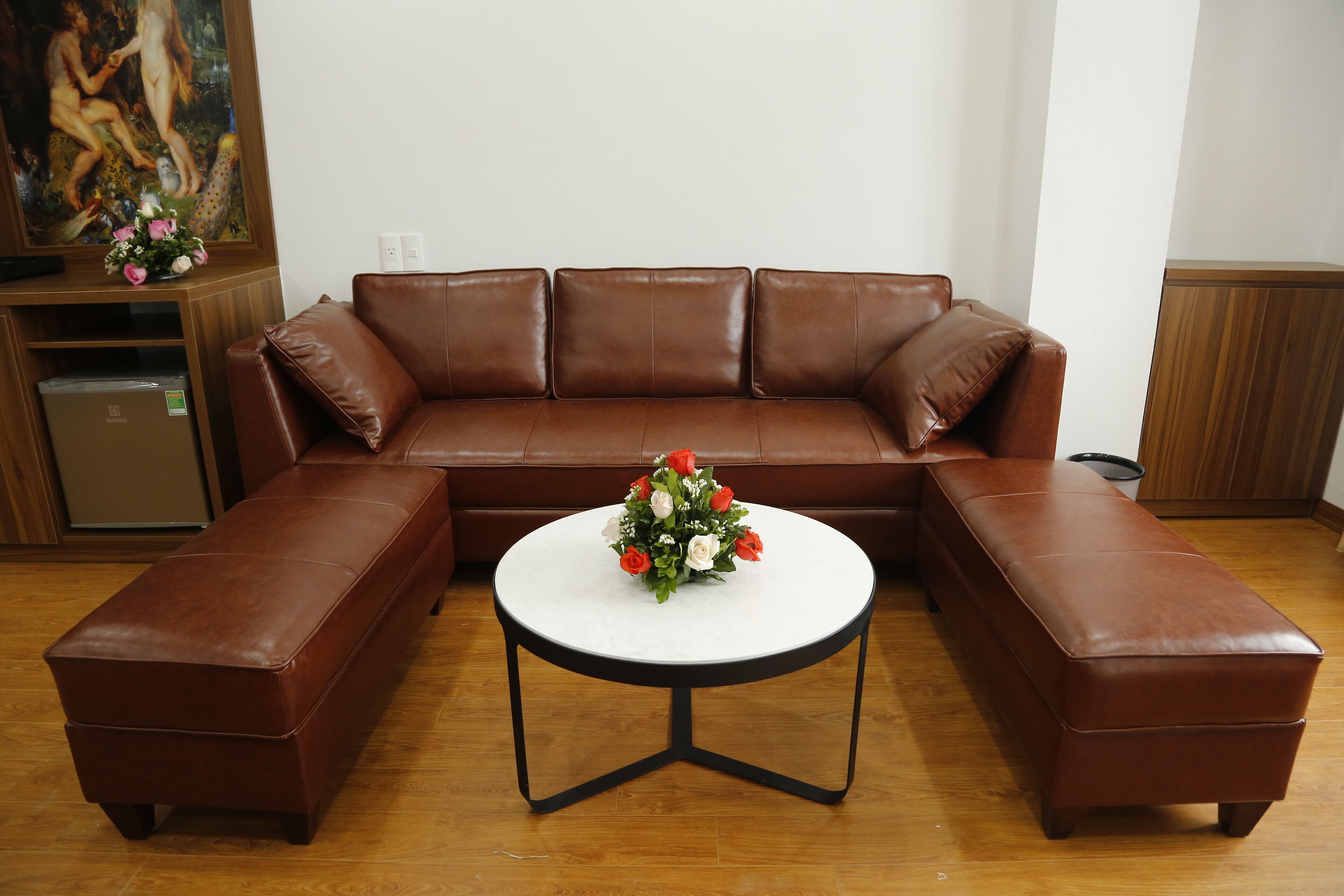 A brown leather sectional sofa with a white round table in the center, a bouquet of flowers, and a painting on the wall.