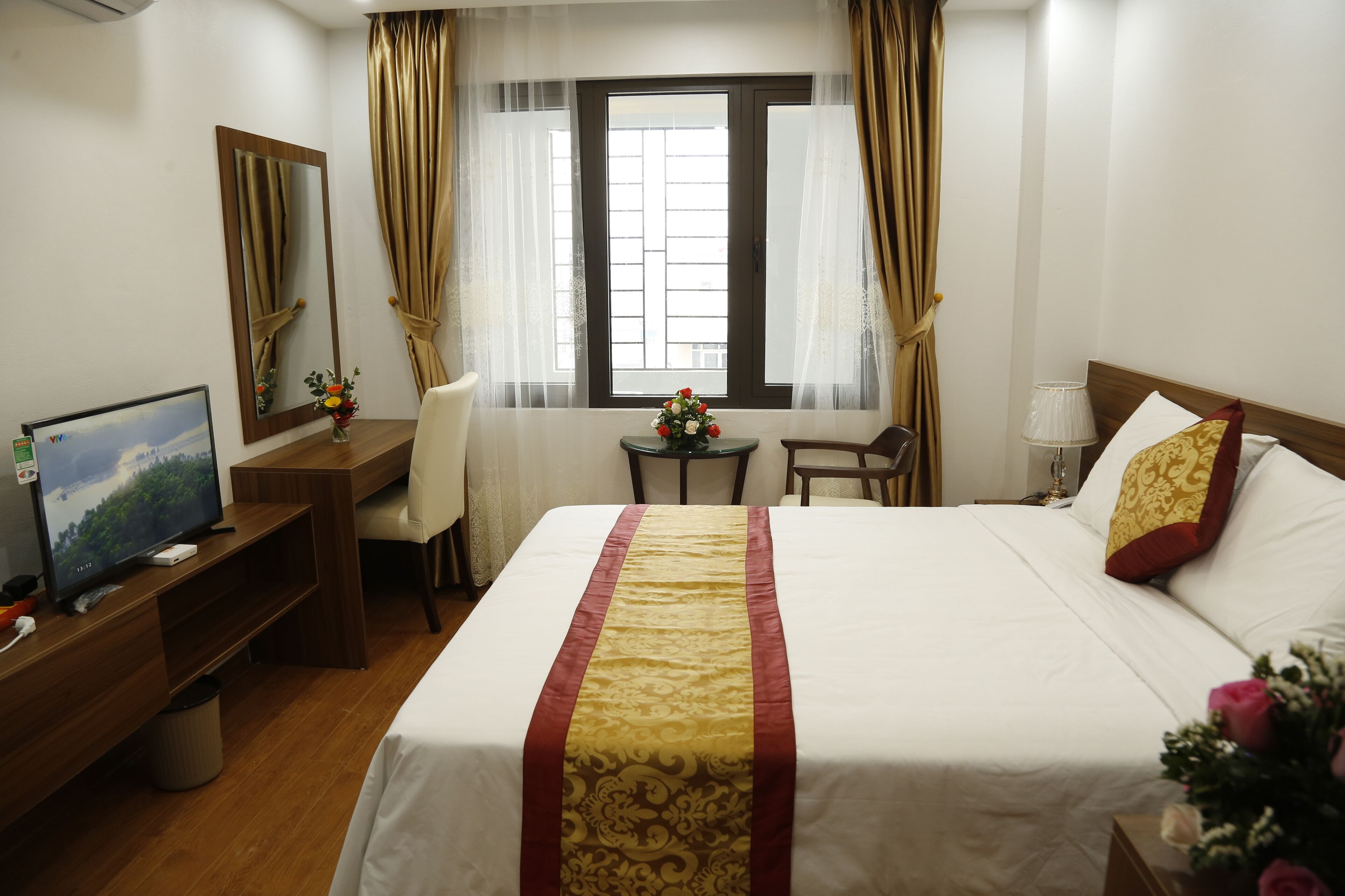 Habitación de hotel con una cama, un escritorio con computadora, una silla, una mesita con un florero y una ventana con cortinas.