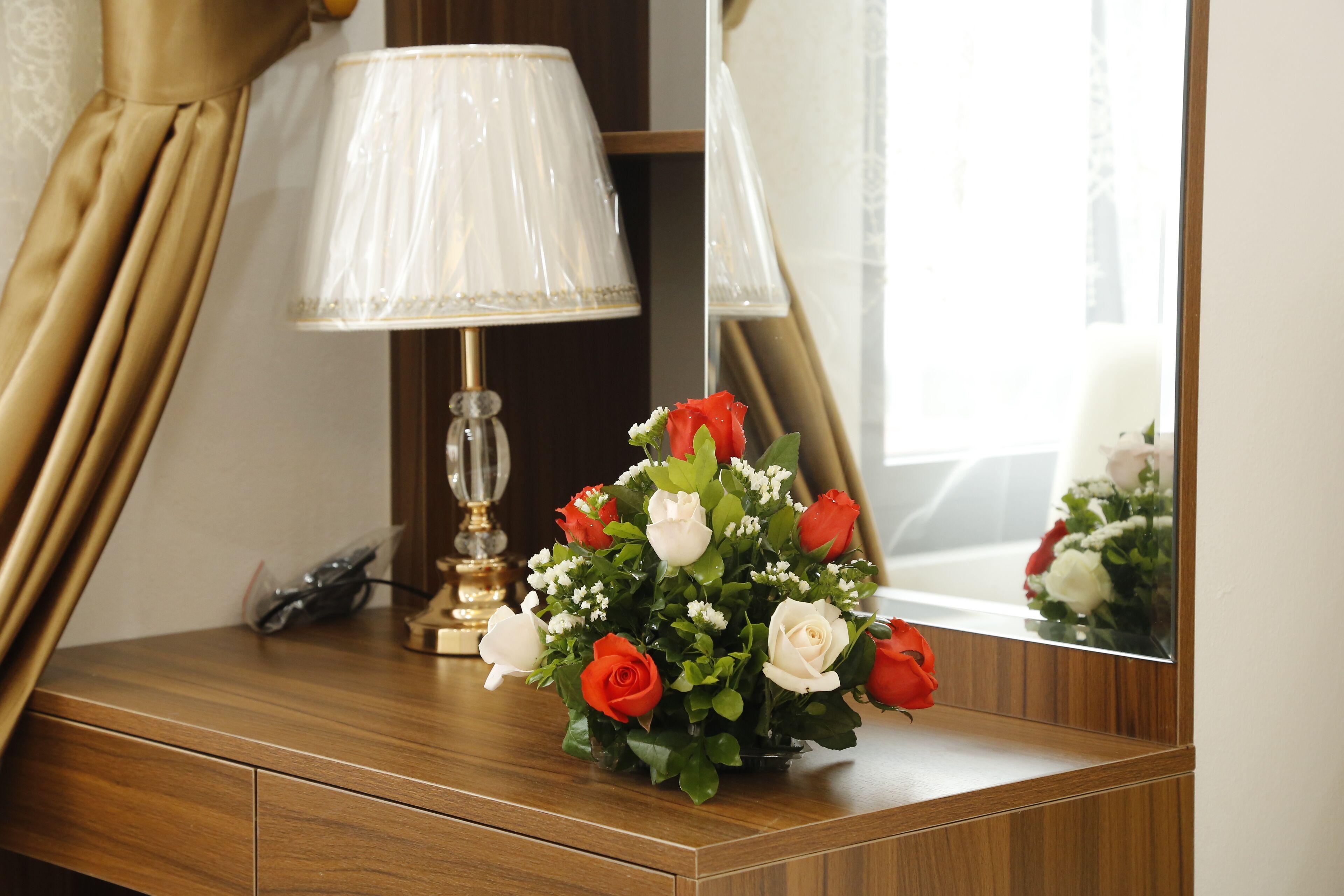 Une commode en bois ornée d’un vase contenant des roses rouges et blanches, une lampe et un miroir qui reflète la vue par la fenêtre.